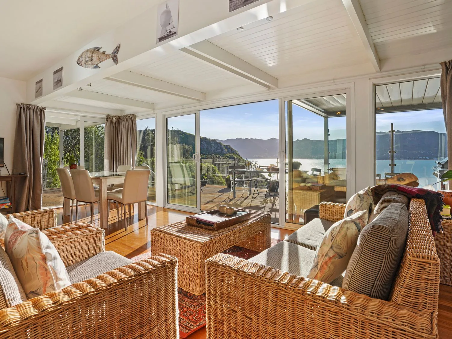 Una sala de estar con muebles de mimbre y una mesa de comedor cerca de grandes puertas correderas de cristal, con vistas a un balcón y a un paisaje montañoso en Kaioruru View - Diamond Harbour Holiday Home, en Diamond Harbour