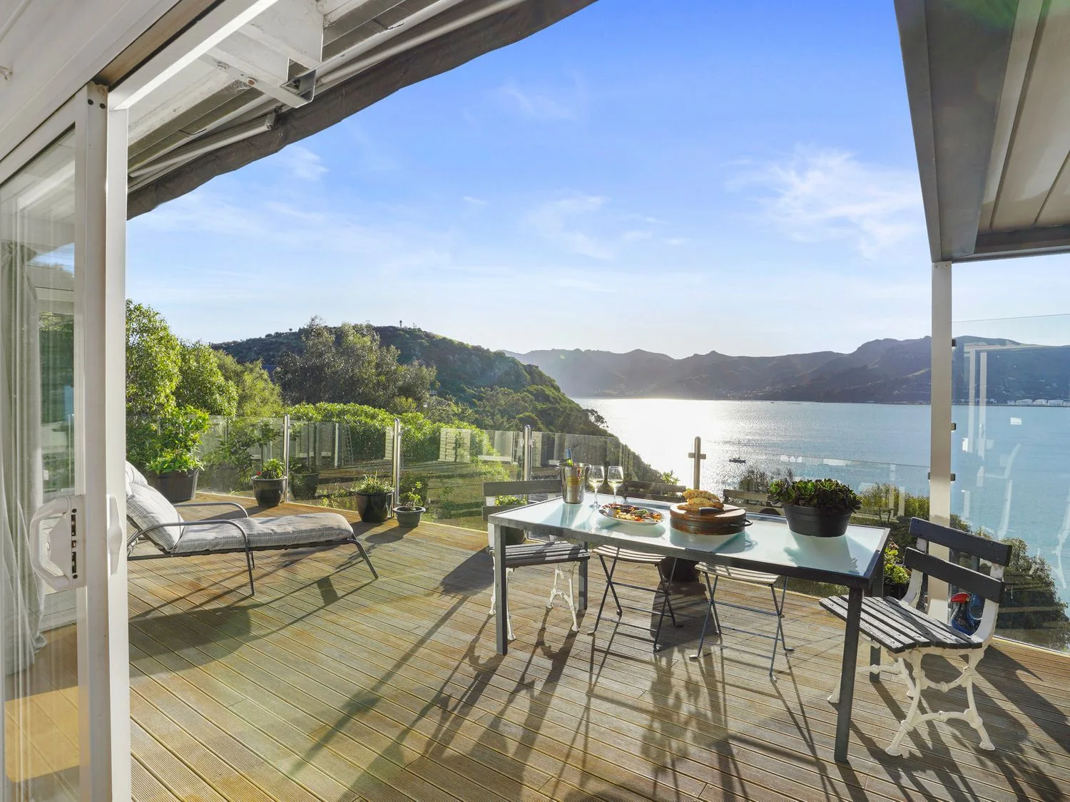 Una terraza de madera con una mesa de cristal, sillas, plantas y una tumbona, con vistas a una bahía con colinas en Kaioruru View - Diamond Harbour Holiday Home, en Diamond Harbour