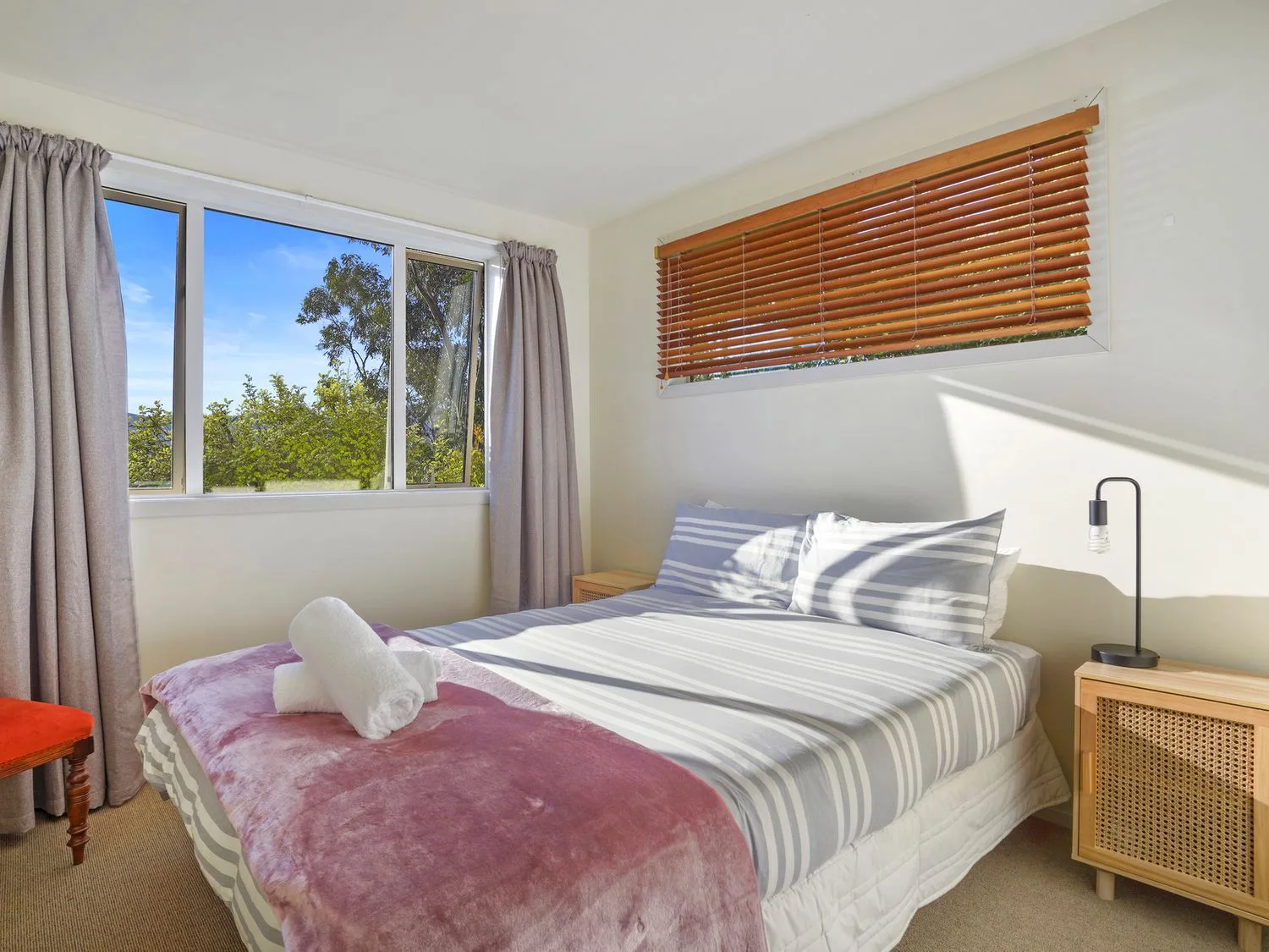 Un dormitorio con una cama vestida con ropa de cama a rayas y una manta rosa bajo ventanas con cortinas y persianas.