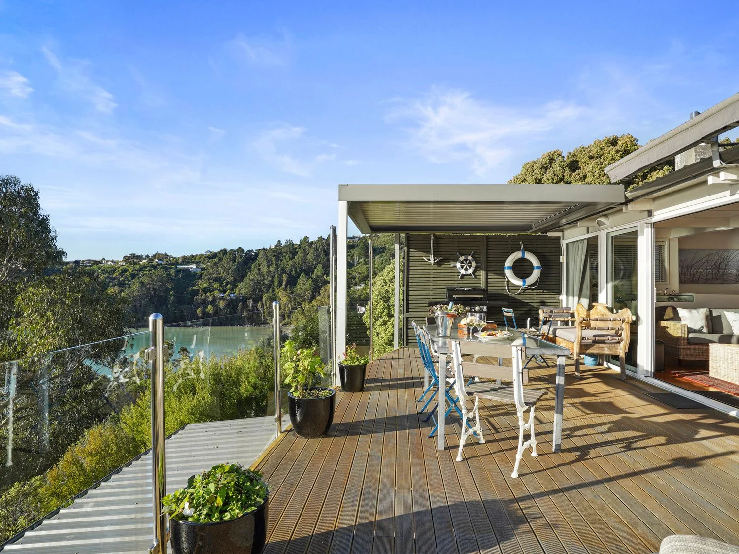 Una terraza de madera con mesa de comedor y sillas, plantas, barandilla de cristal y vistas a los árboles y al agua en Kaioruru View Diamond Harbour Holiday Home, en Diamond Harbour