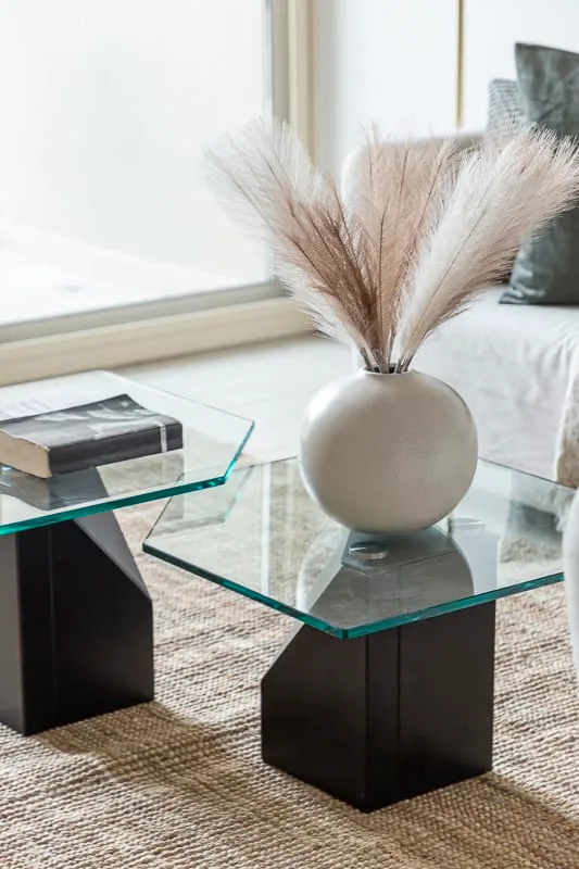 A glass coffee table with a vase of dried grasses sits beside a sofa.
