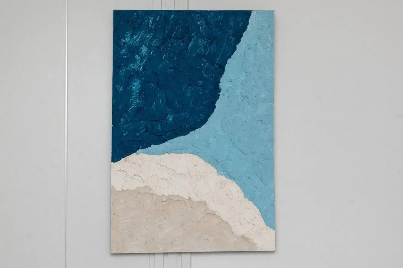 Una pintura abstracta texturizada en azul y crema cuelga de una pared clara.