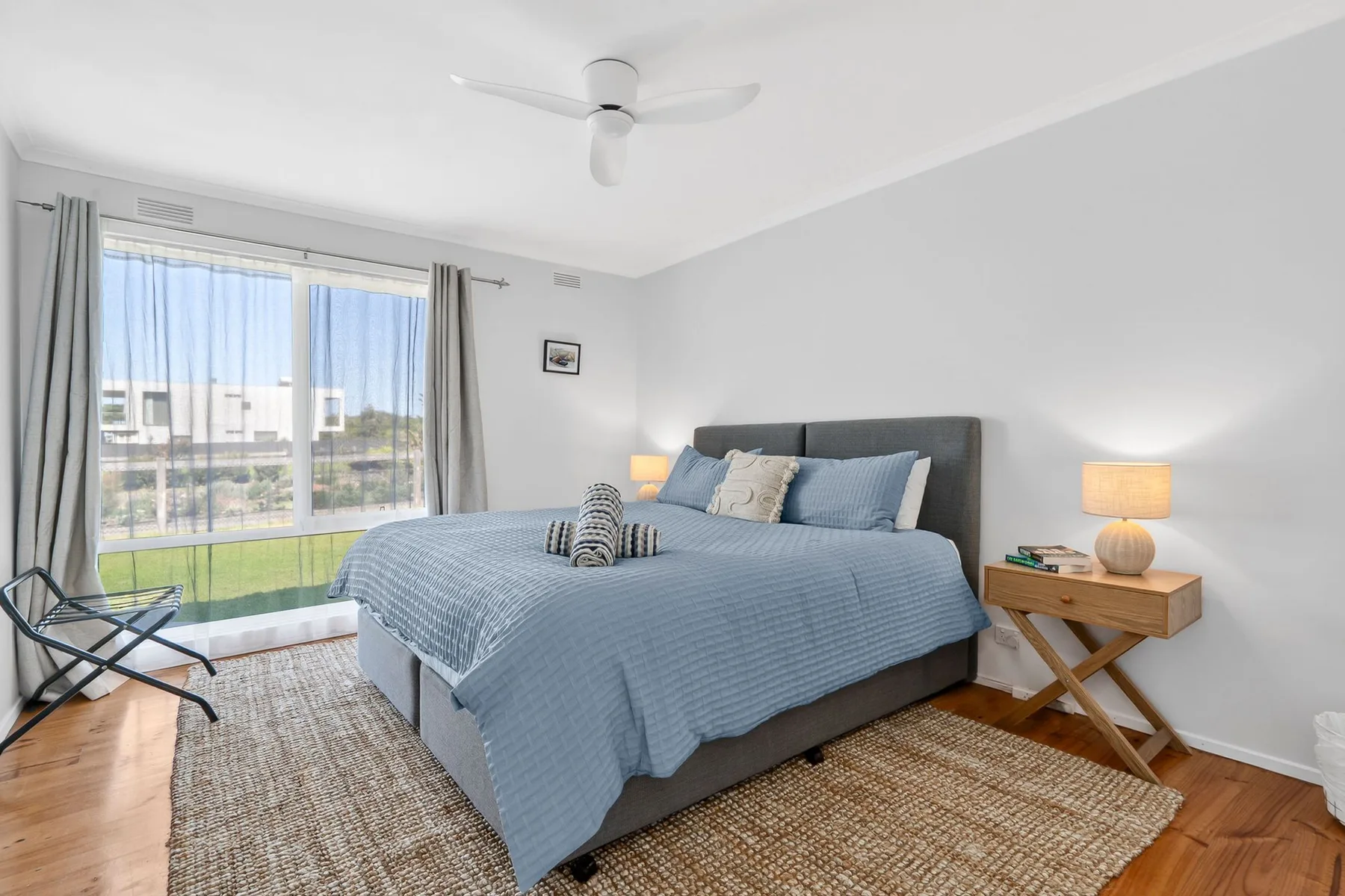 A bright bedroom with a blue bed, bedside tables, and a large window.