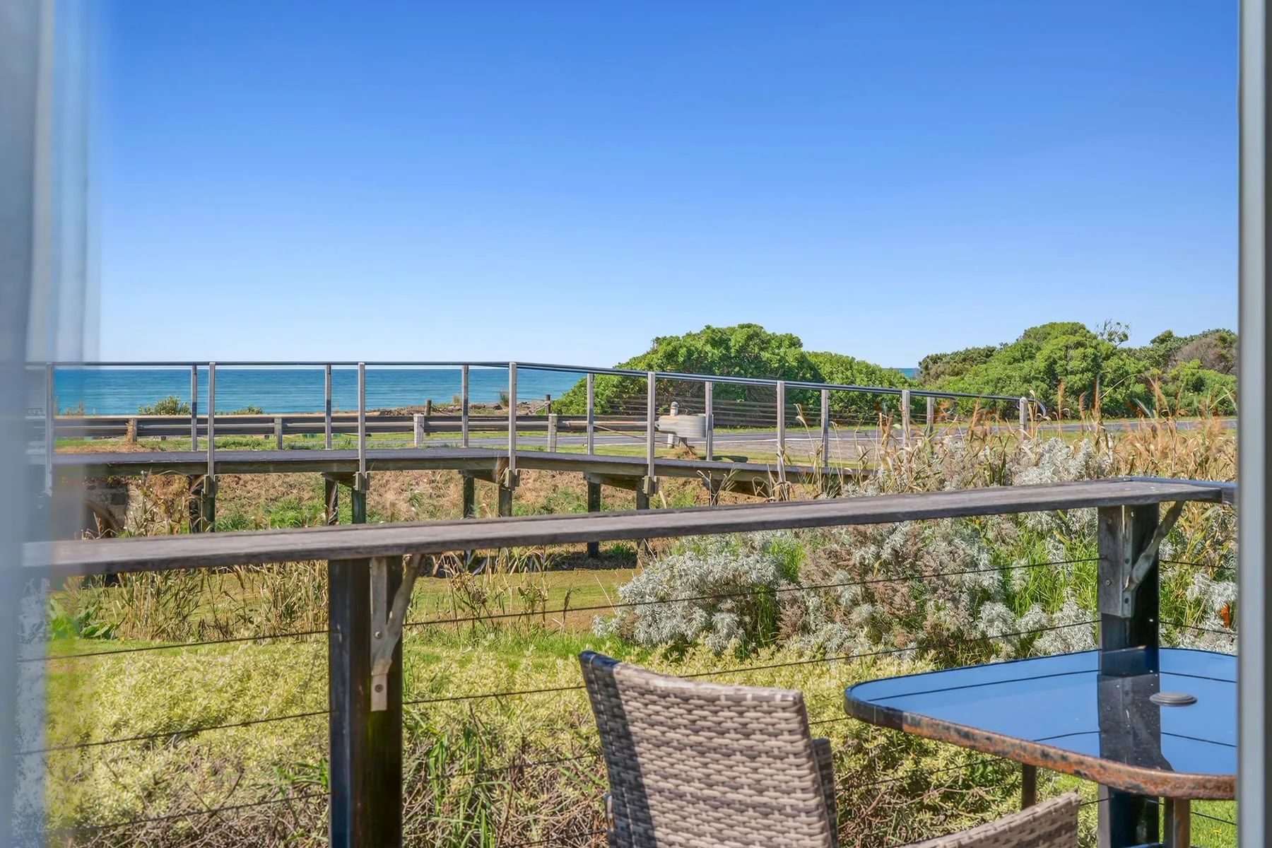 A balcony overlooks green coastal vegetation and the ocean beneath a clear blue sky.