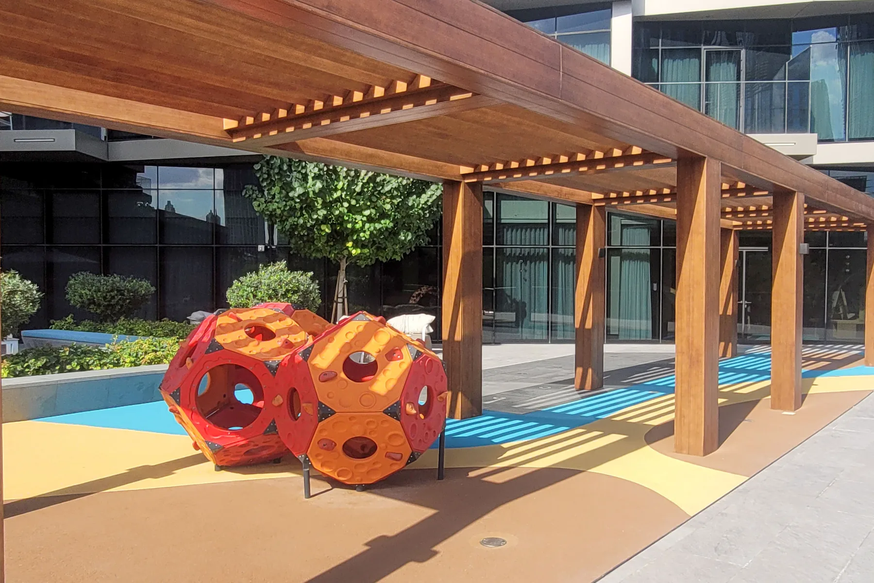 A colorful playground structure sits beneath a wooden pergola beside a modern building.
