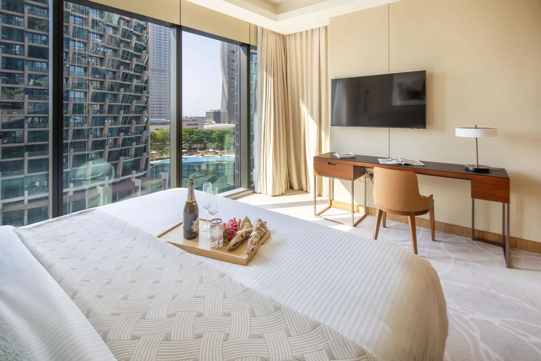 A bright bedroom with a bed, desk, and large windows overlooking nearby buildings.