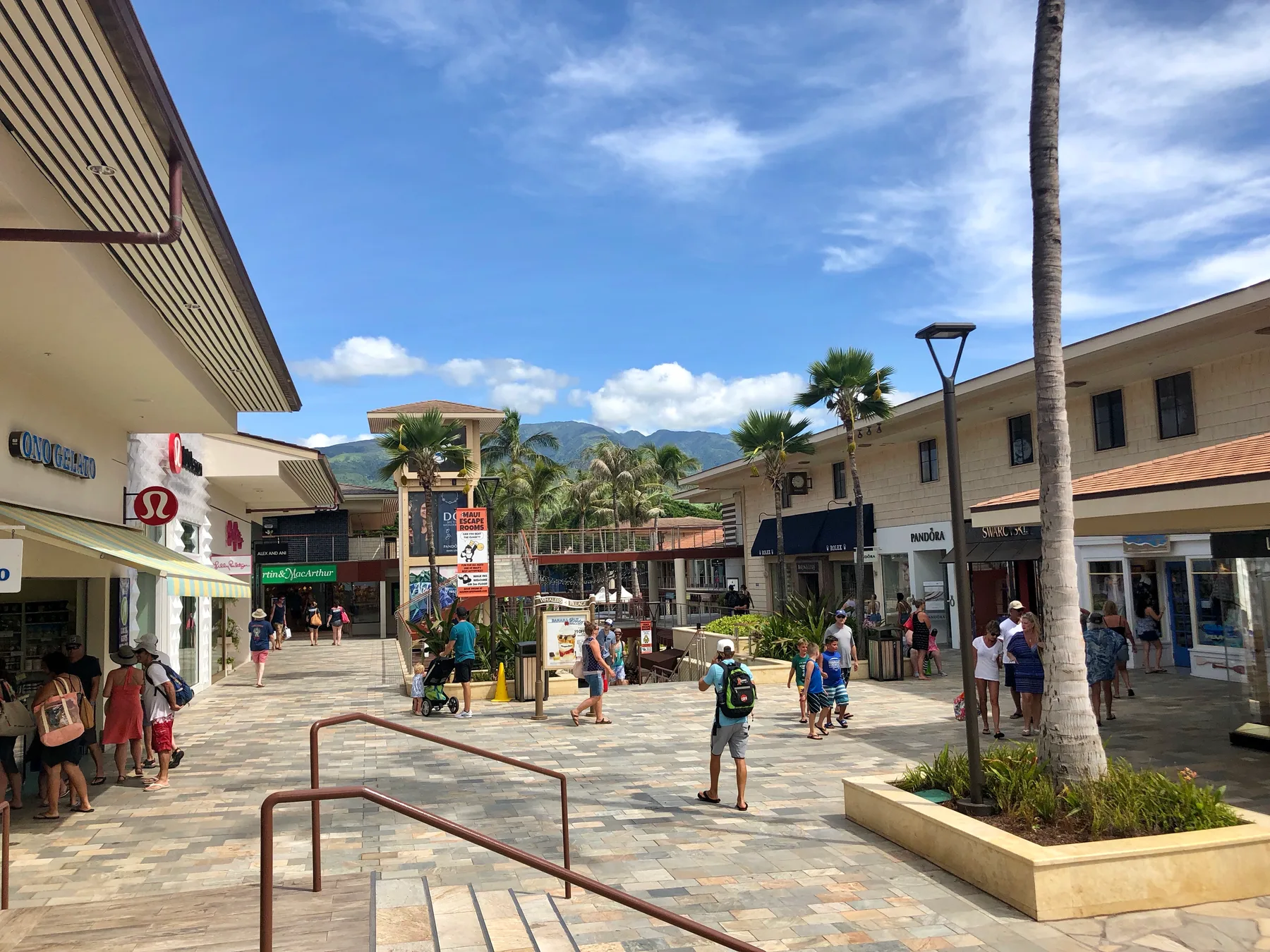 Una plaza comercial al aire libre con tiendas, palmeras y montañas a lo lejos.
