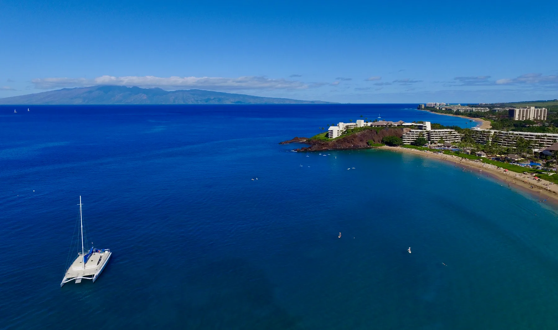 Una bahía costera con un catamarán mar adentro y una playa de arena junto a la costa.