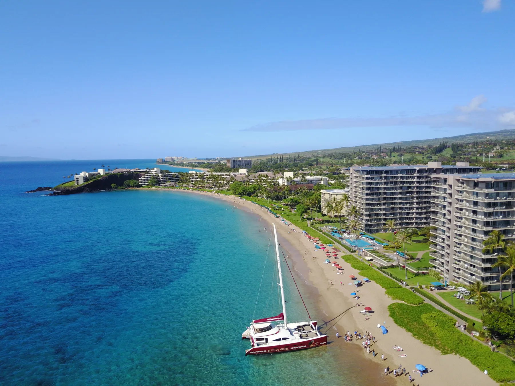 Vista aérea de una playa costera con agua turquesa, un catamarán y edificios frente al mar.