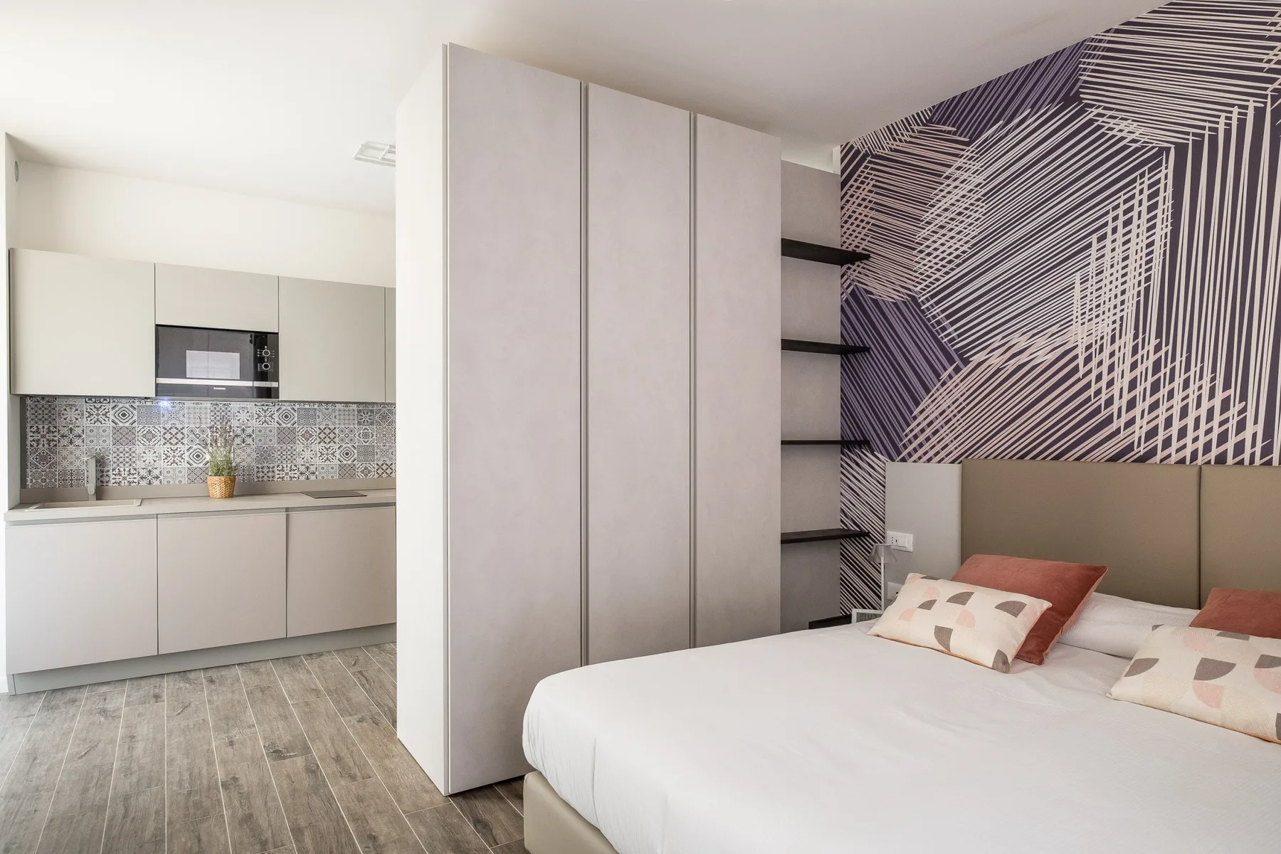 A bedroom with a white bed beside a compact kitchen and patterned wall.