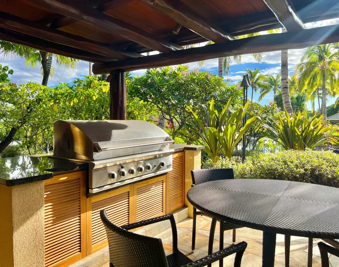 Un patio cubierto tiene una parrilla incorporada, mesa y asientos entre vegetación tropical.