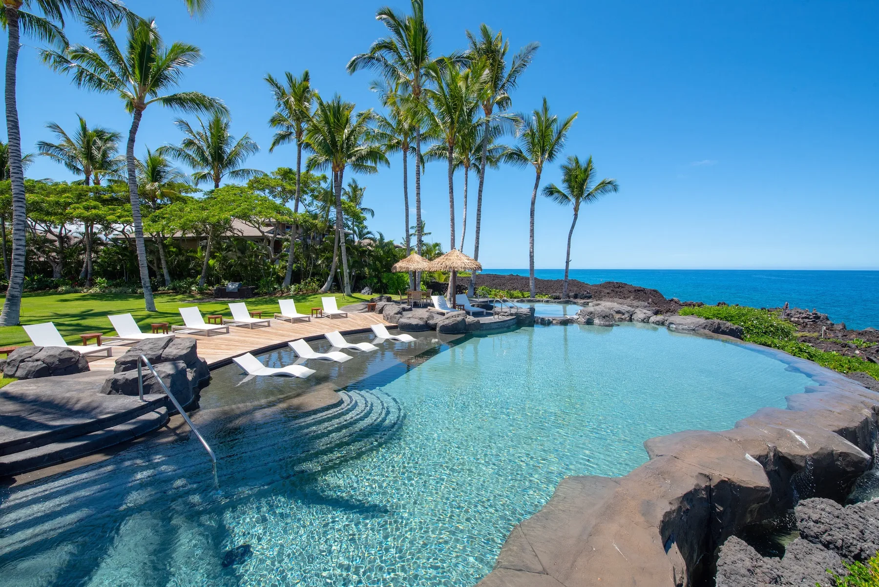 Una piscina tropical junto al océano, con palmeras y tumbonas junto al borde.
