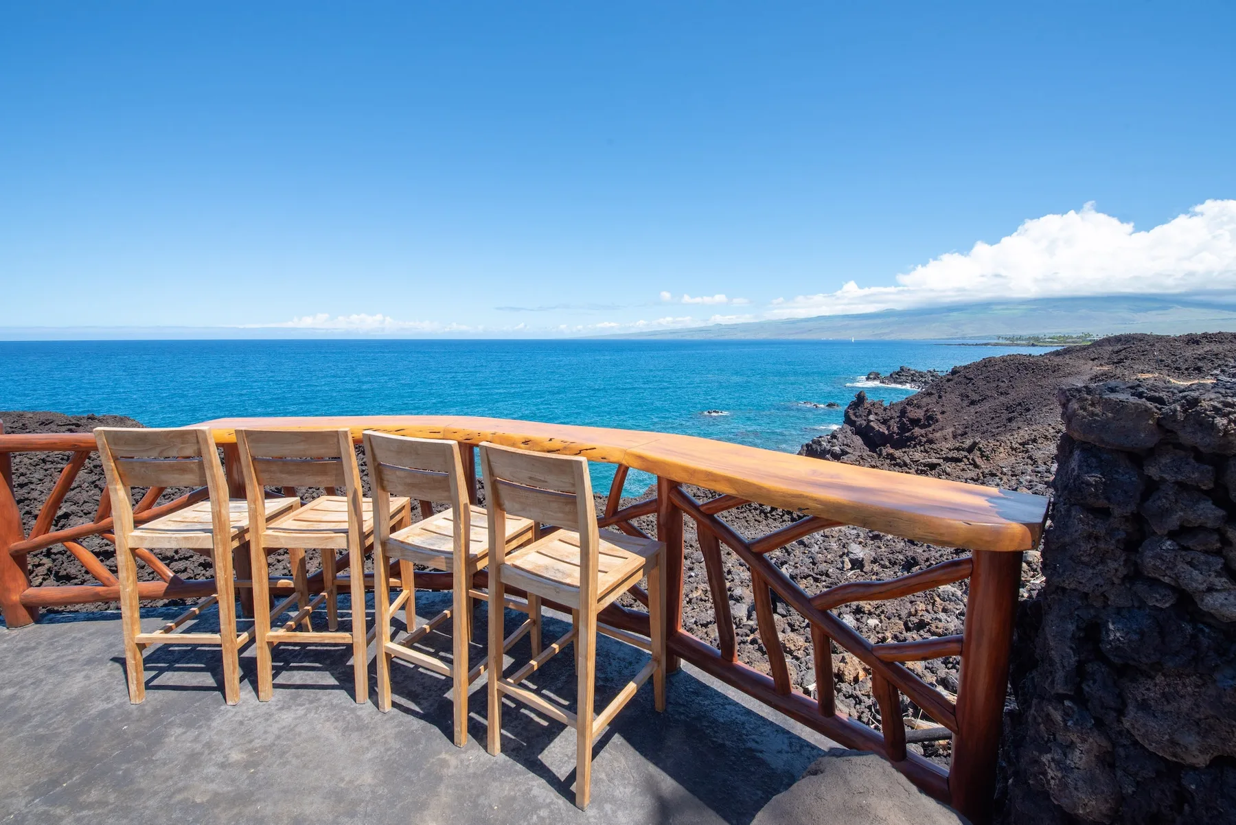 Una terraza junto al mar con asientos de madera da a aguas azules y una costa rocosa.
