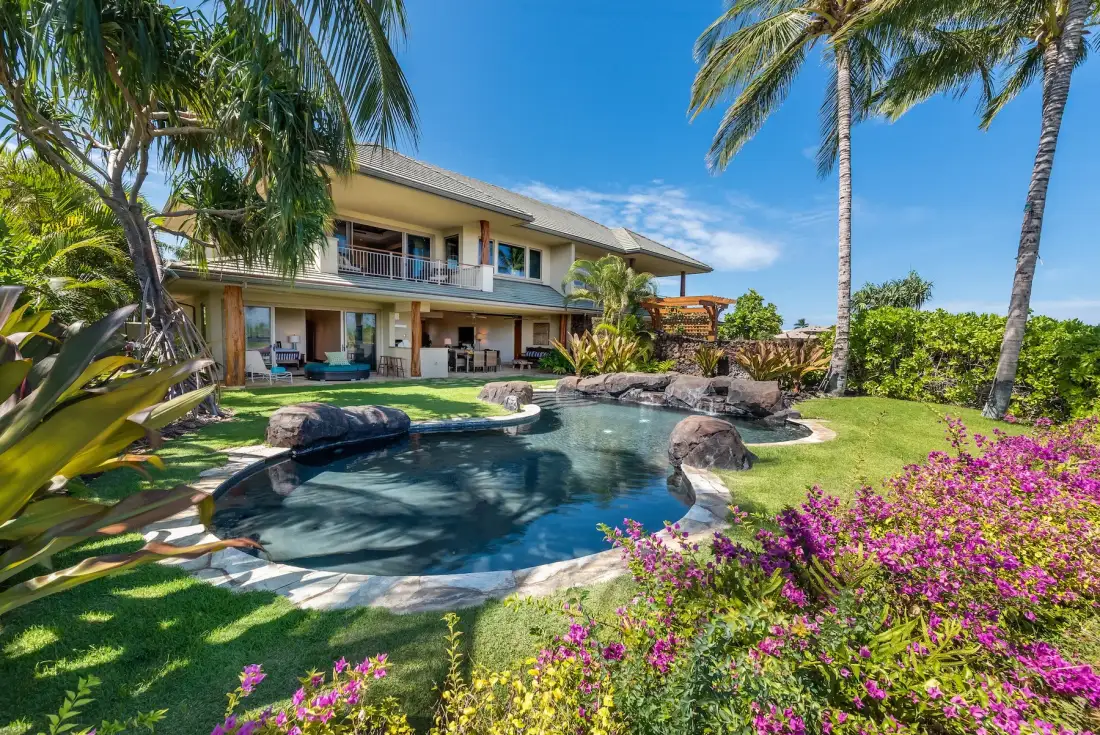 Una casa tropical da a una piscina curva rodeada de jardines y palmeras.