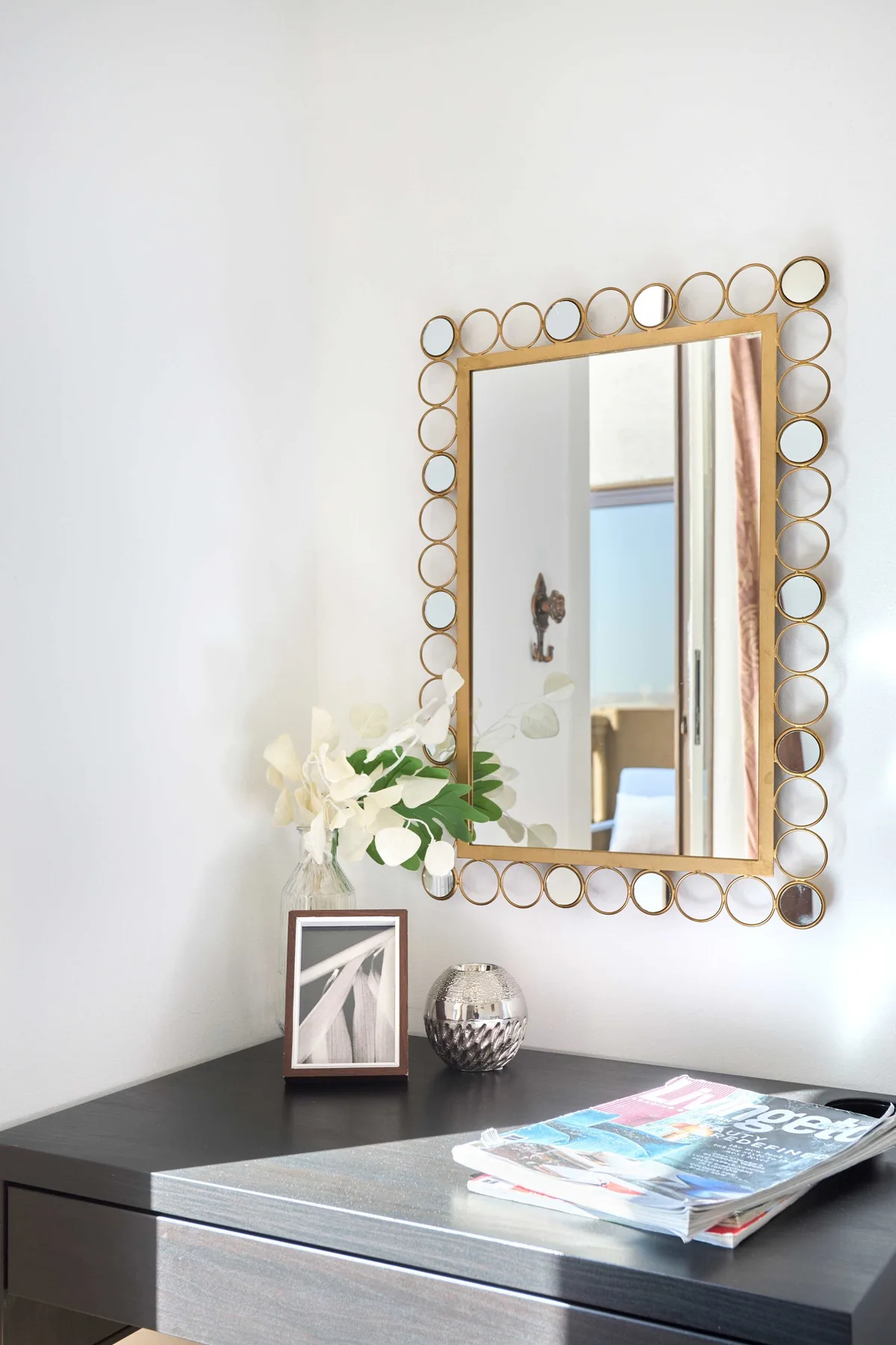 Una consola oscura está bajo un espejo decorativo, con flores y revistas encima.