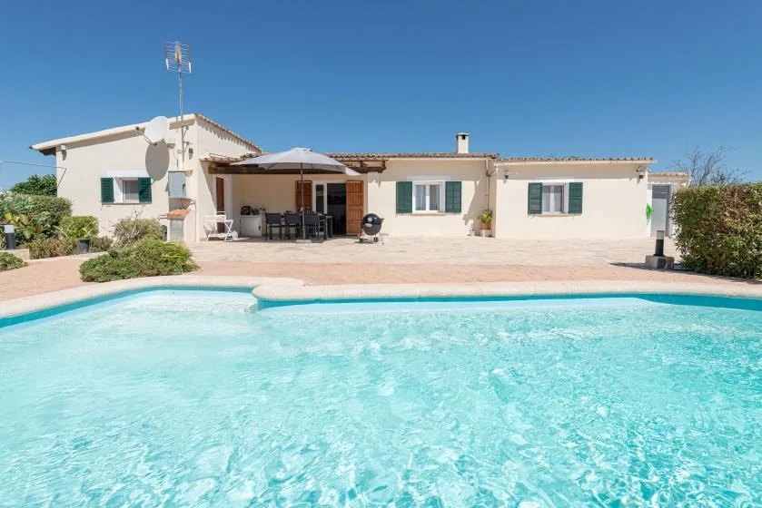 Una piscina frente a una casa de una planta con un patio sombreado.