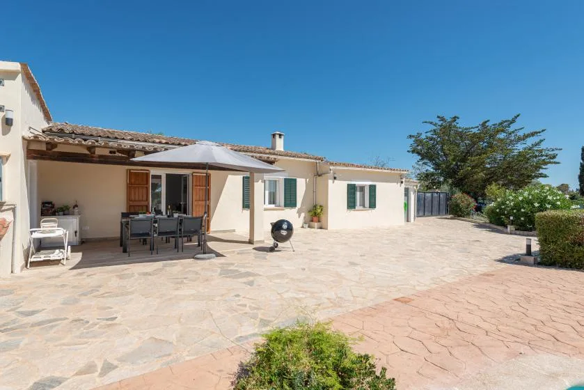 A house opens onto a spacious stone-paved patio with outdoor seating and an umbrella.