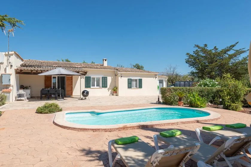 Un patio soleado con piscina, asientos al aire libre y una casa baja.