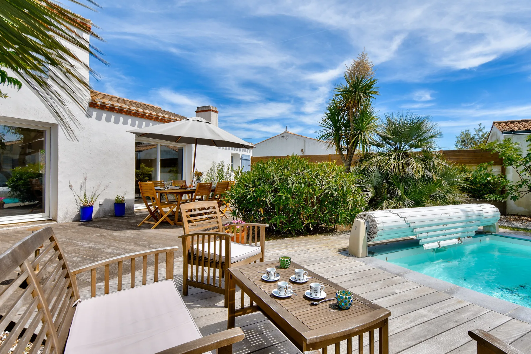 A sunny outdoor patio with wooden seating, a table, and a swimming pool.