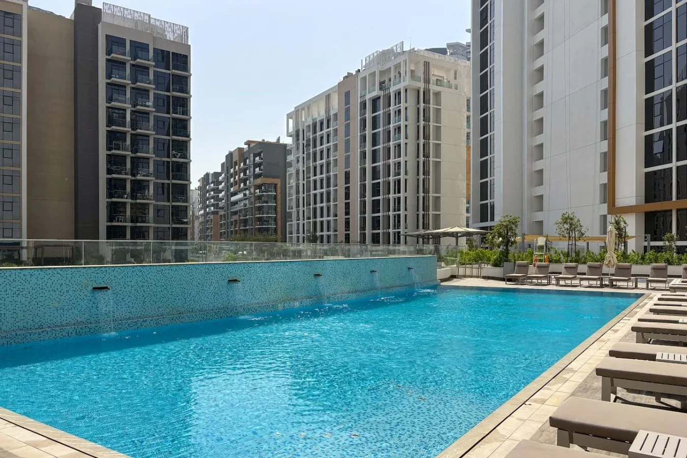 An outdoor swimming pool is surrounded by lounge seating and tall apartment buildings.
