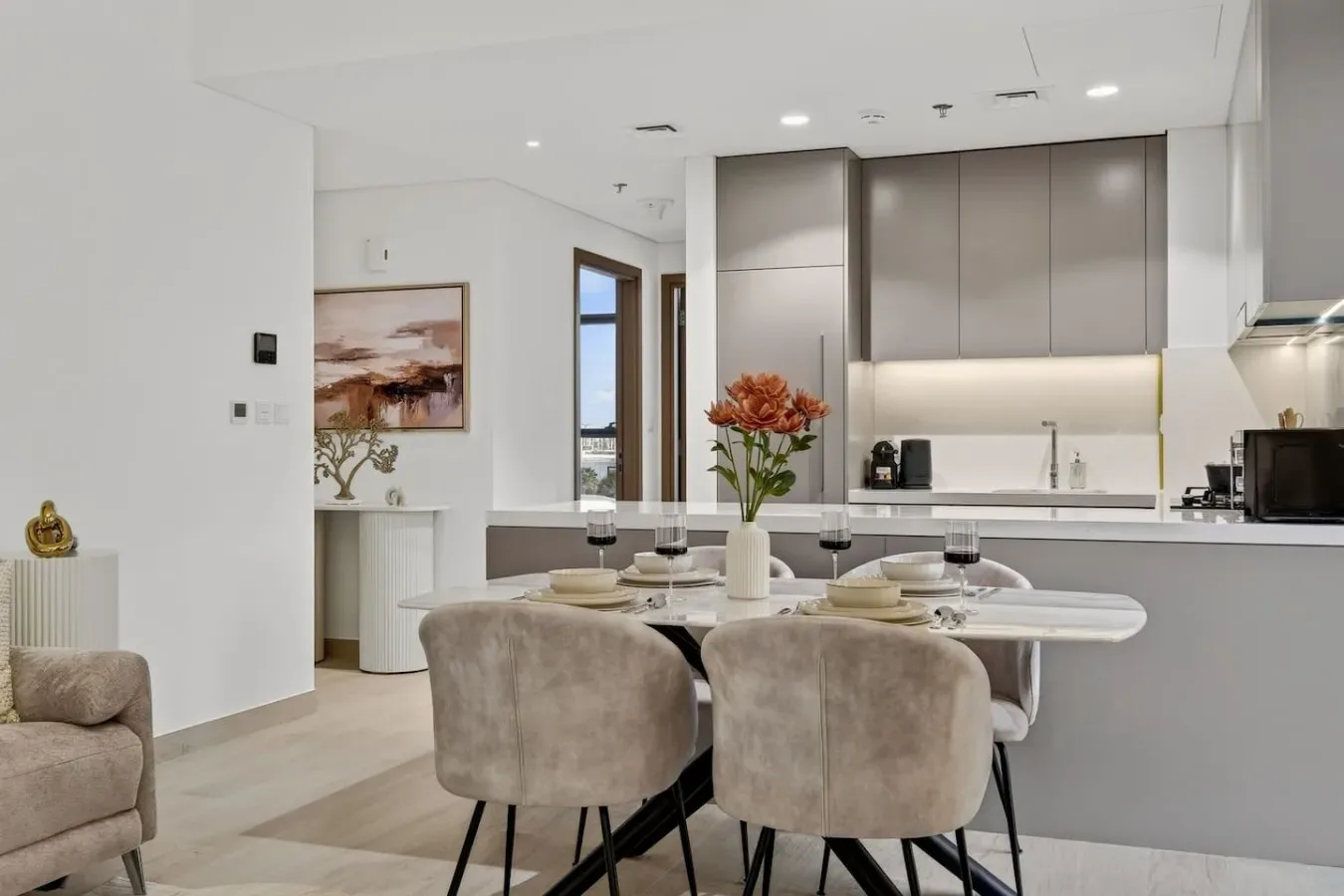 A modern dining area beside an open kitchen, with a set table and flower arrangement.
