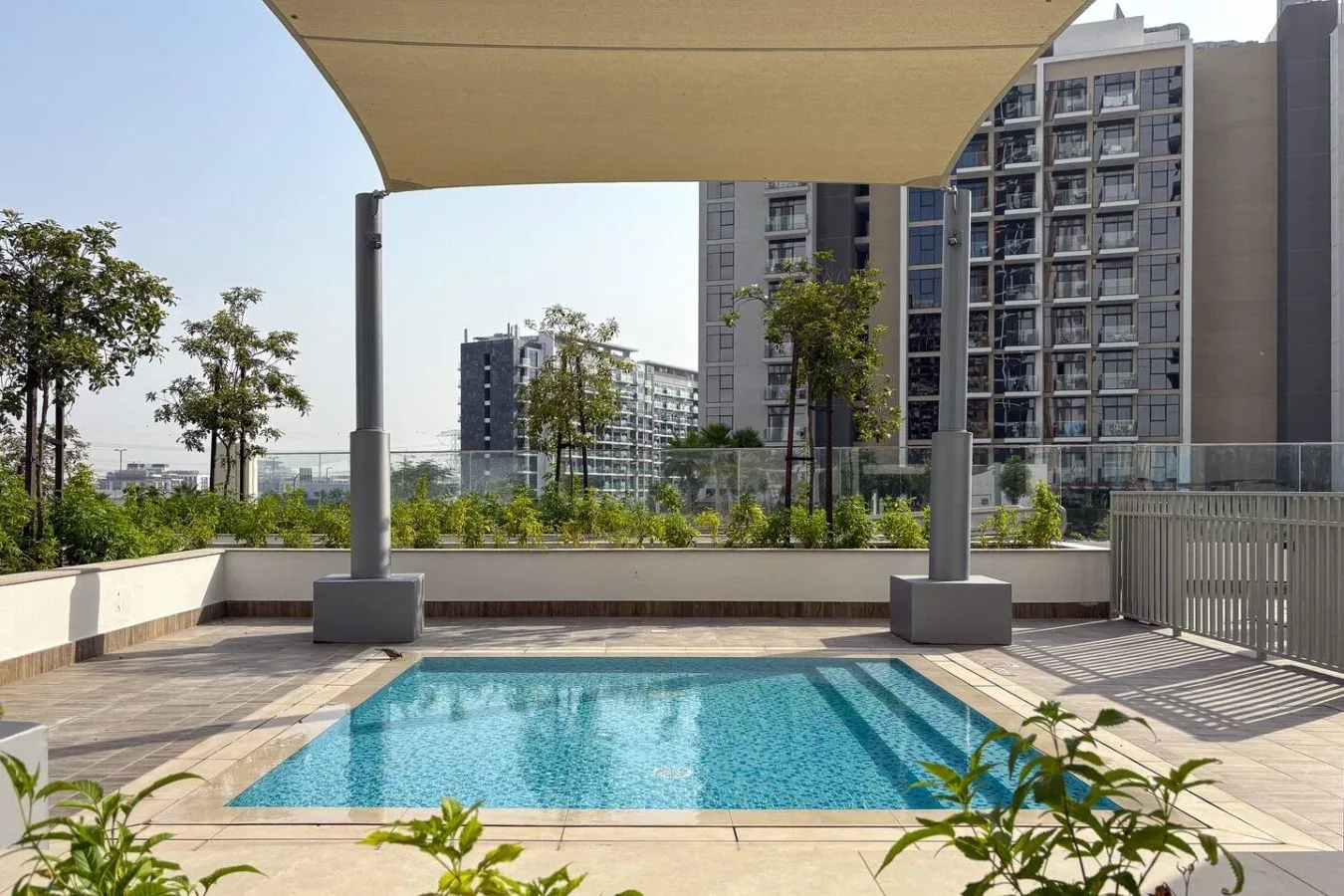 An outdoor swimming pool sits beneath a shade canopy, with apartment buildings in the background.