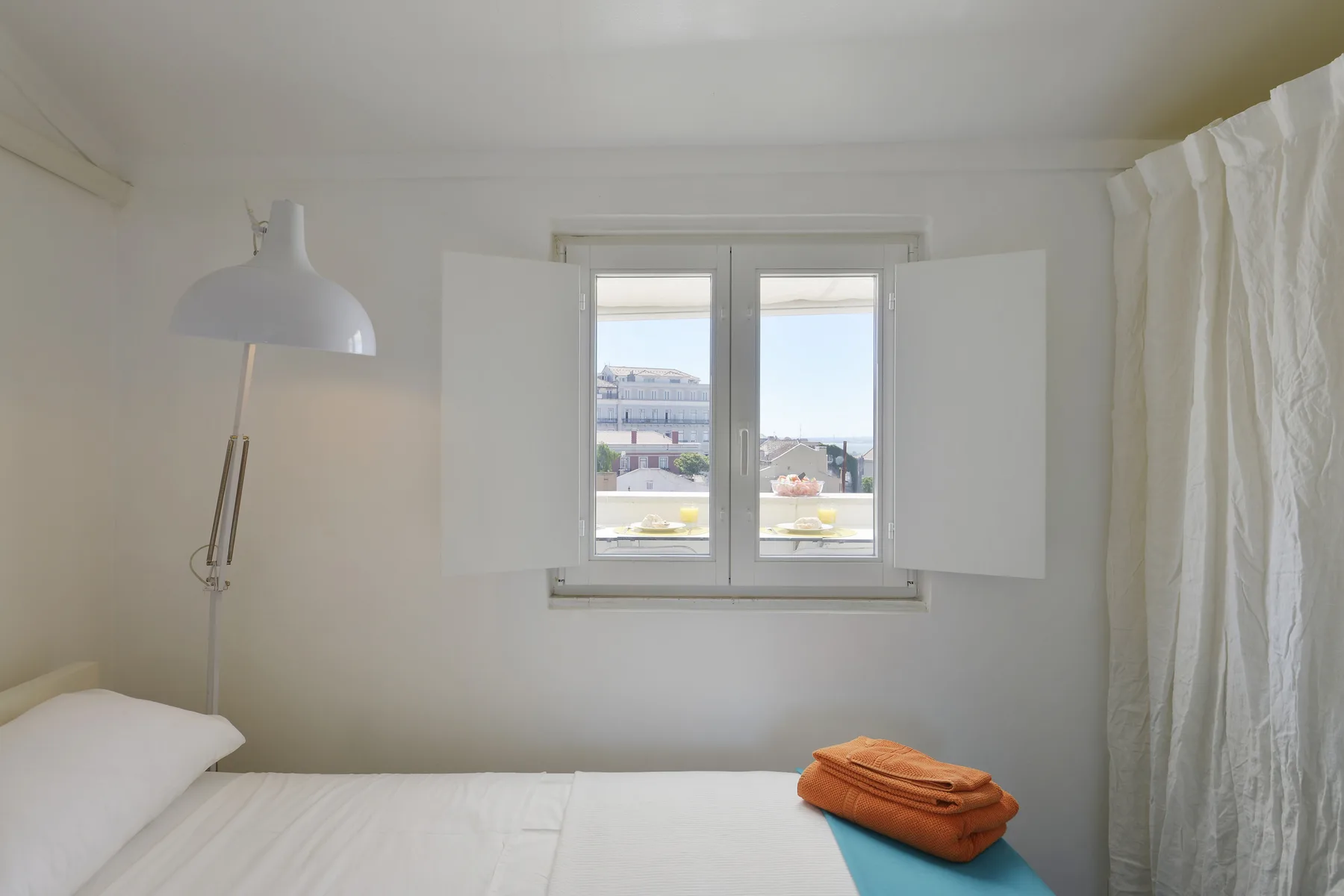 A bright bedroom with a bed, bedside lamp, and a window overlooking rooftops.