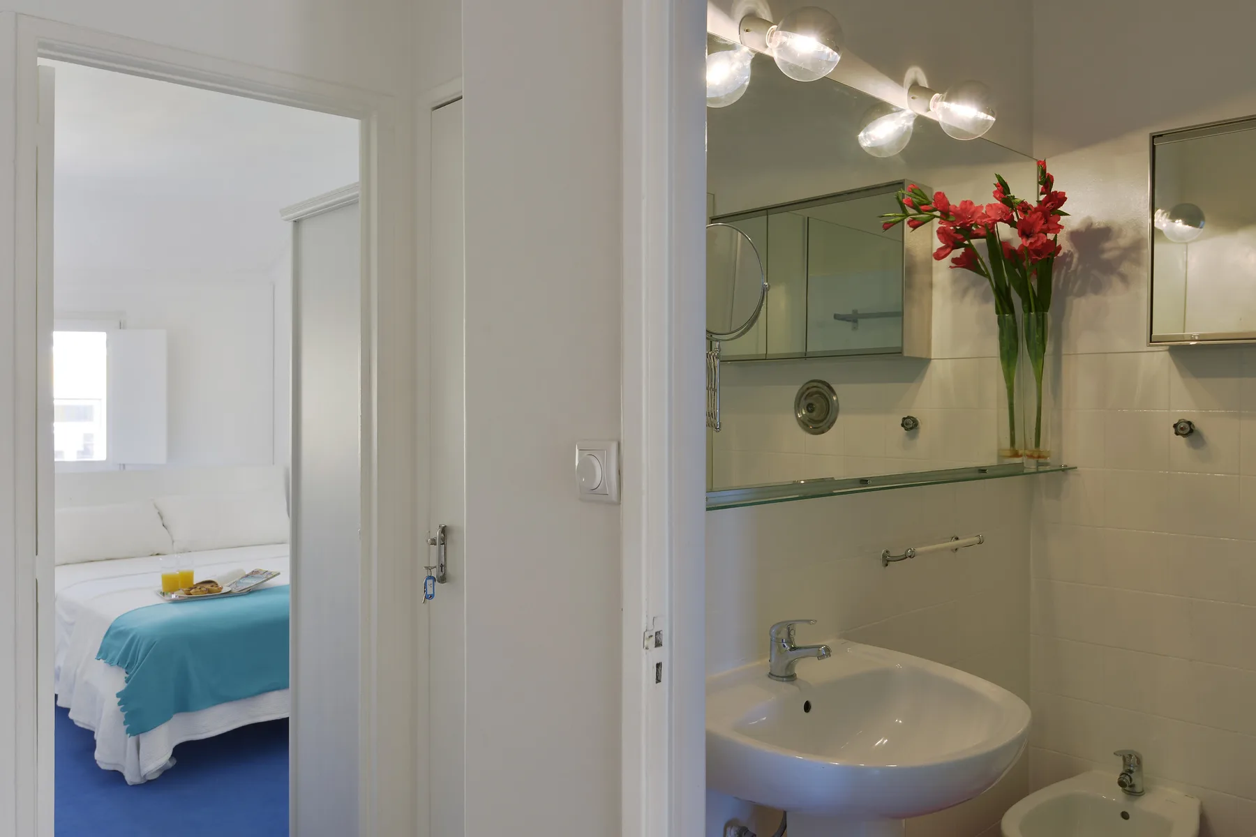 A bedroom with blue bedding is visible through the doorway beside a bathroom sink.