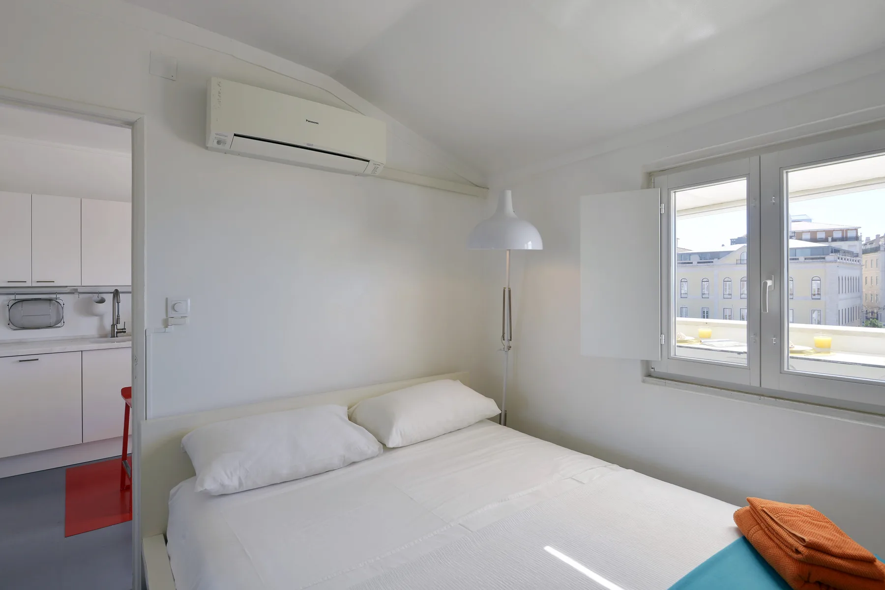 A bright bedroom with a bed and windows overlooking nearby buildings.