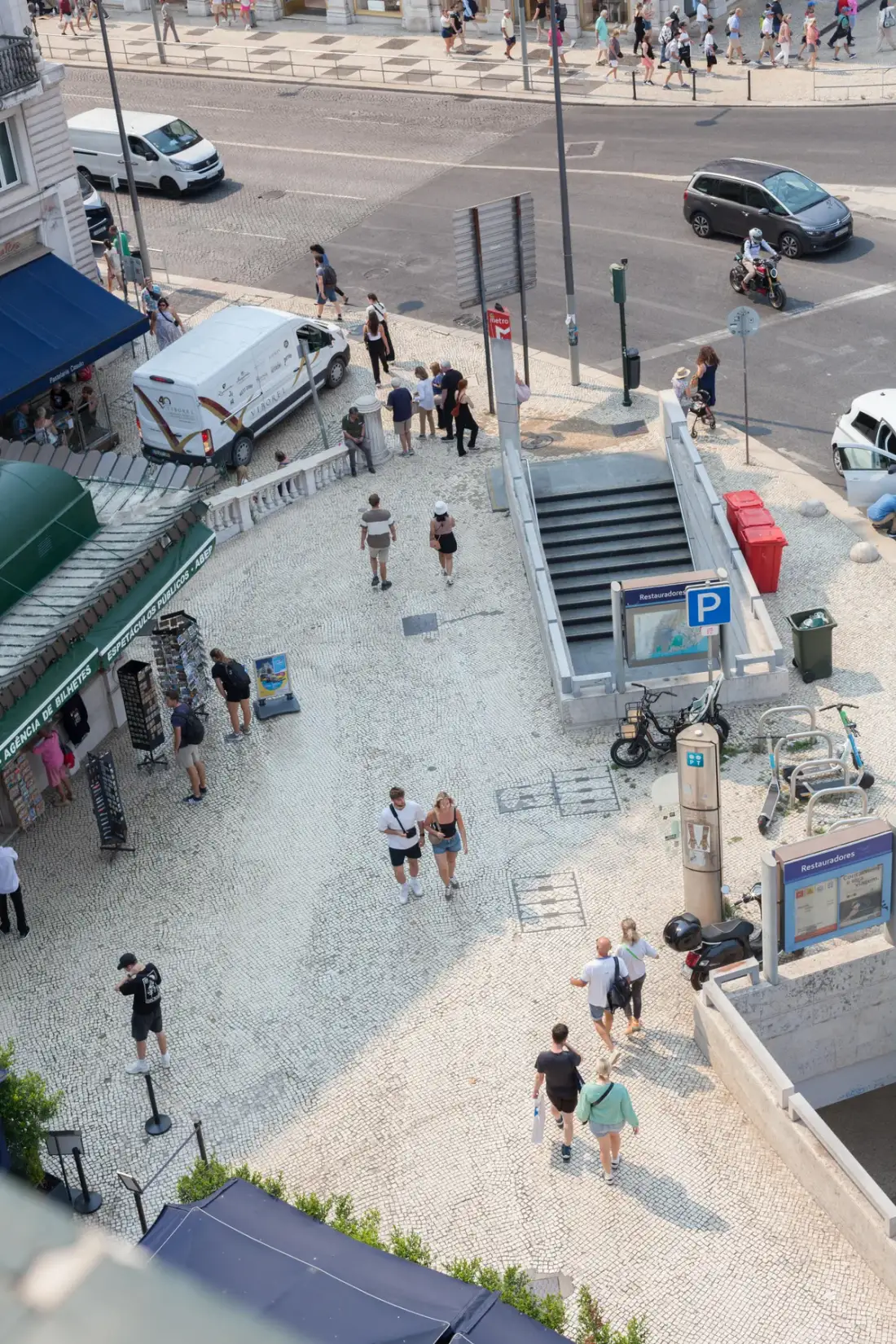 Una plaza peatonal concurrida con personas caminando junto a una calle y vehículos estacionados.