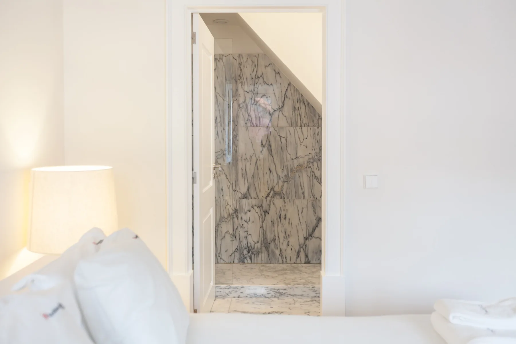 Un dormitorio luminoso con ropa de cama blanca y una puerta abierta a un baño de mármol.