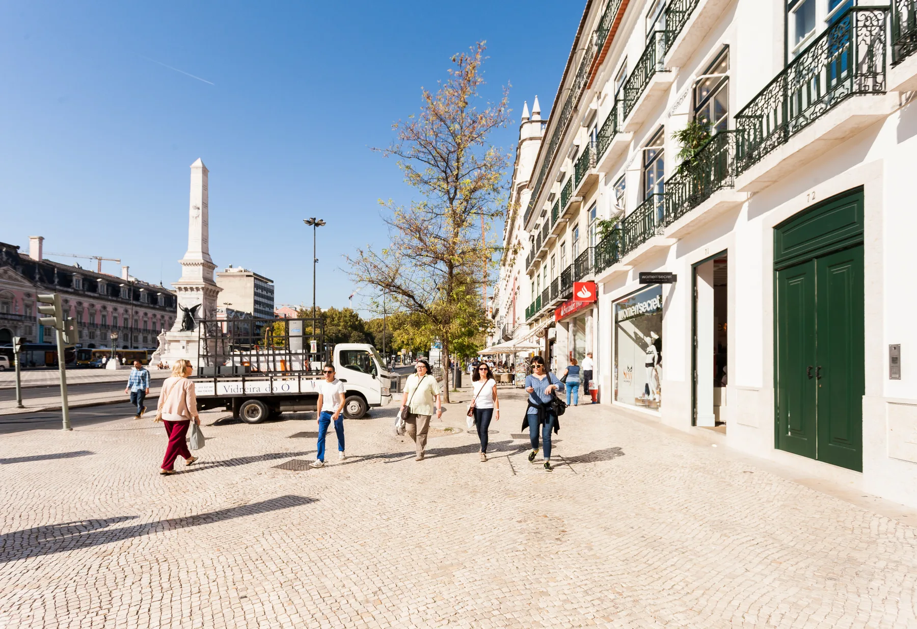 Una plaza soleada con un monumento alto de piedra, comercios y peatones.