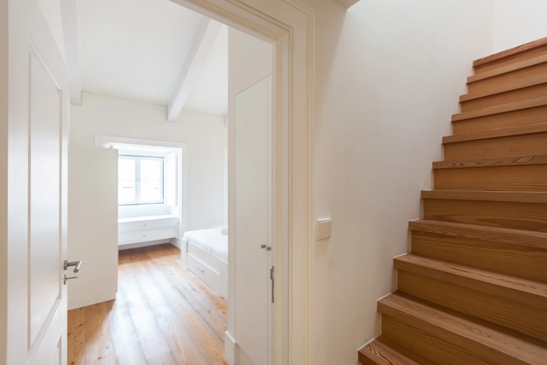 A través de una puerta abierta se ve una habitación luminosa con ventana junto a una escalera de madera.