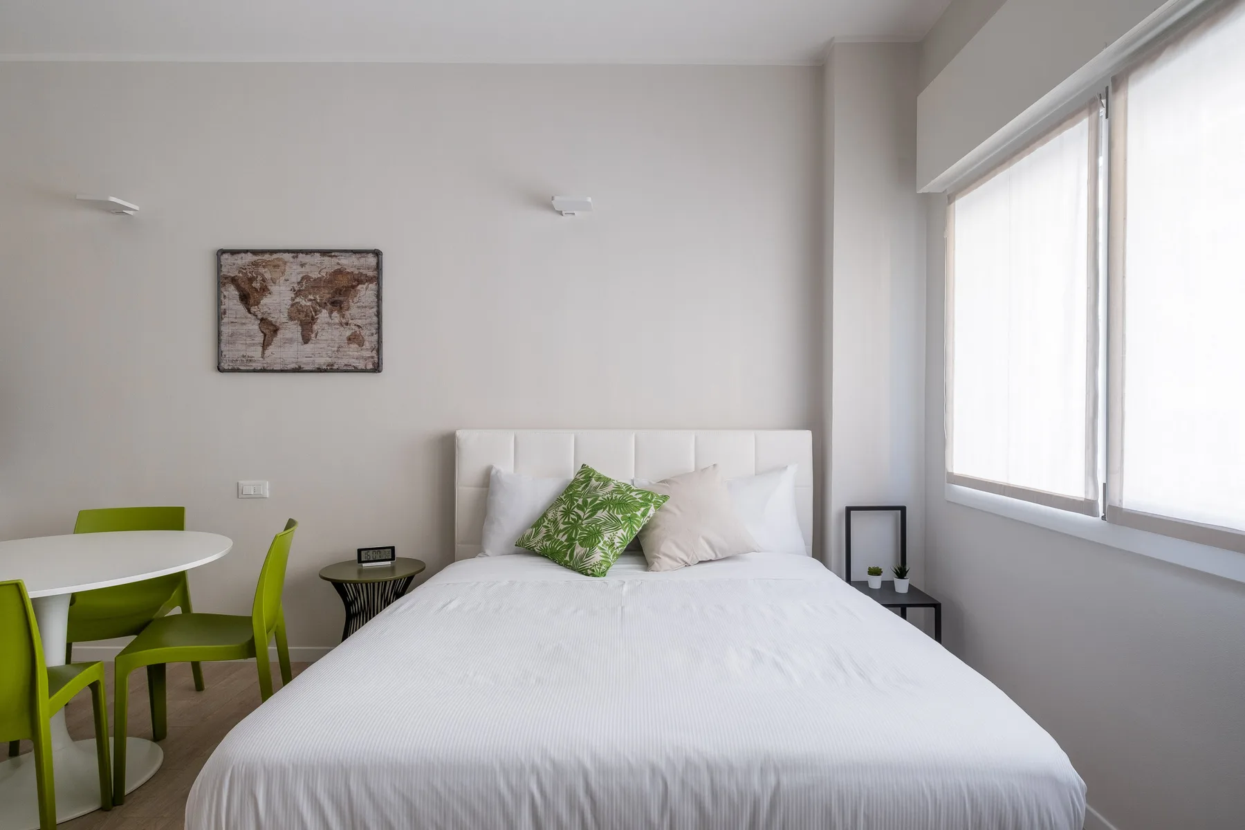 Un dormitorio luminoso con una cama tendida y asientos verdes junto a una mesa pequeña.