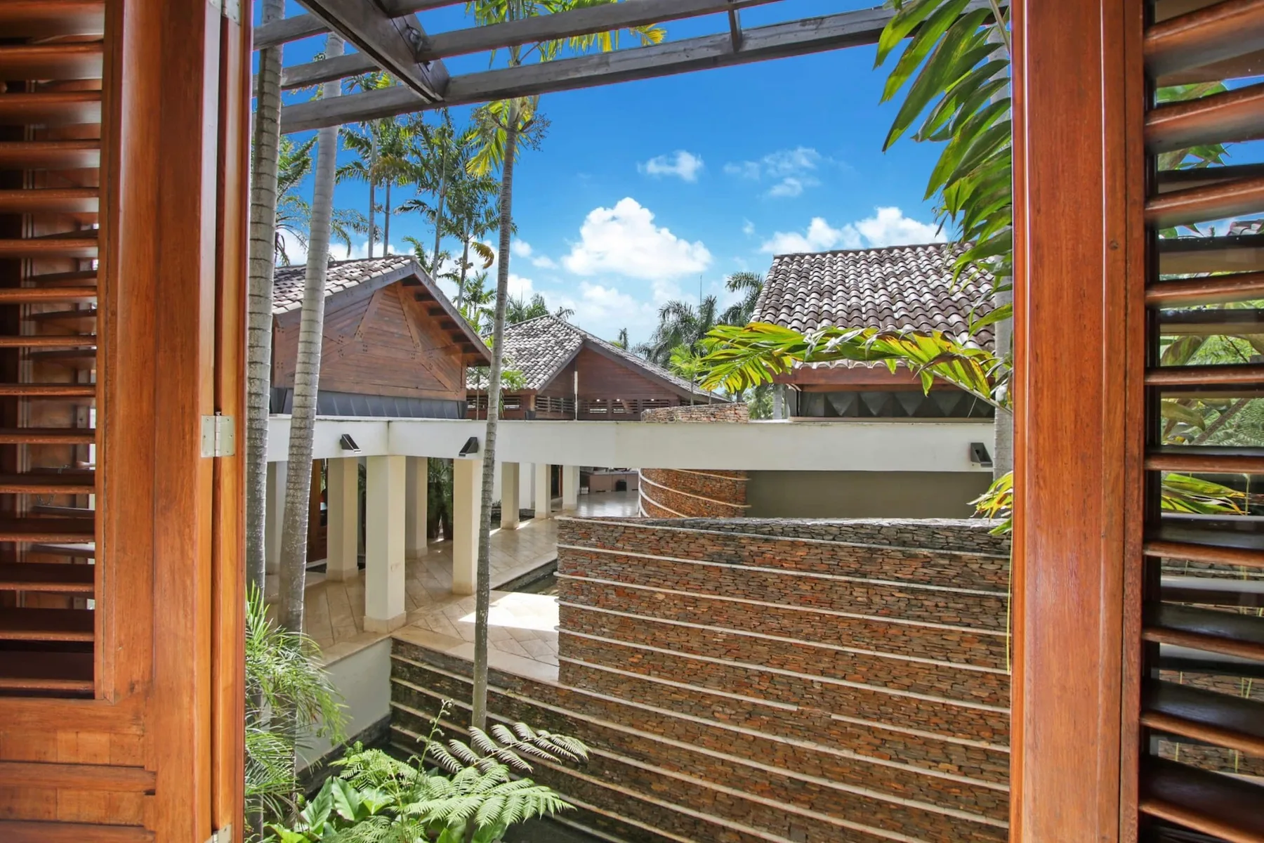 A courtyard view through wooden shutters, with tiled roofs and tropical plants.