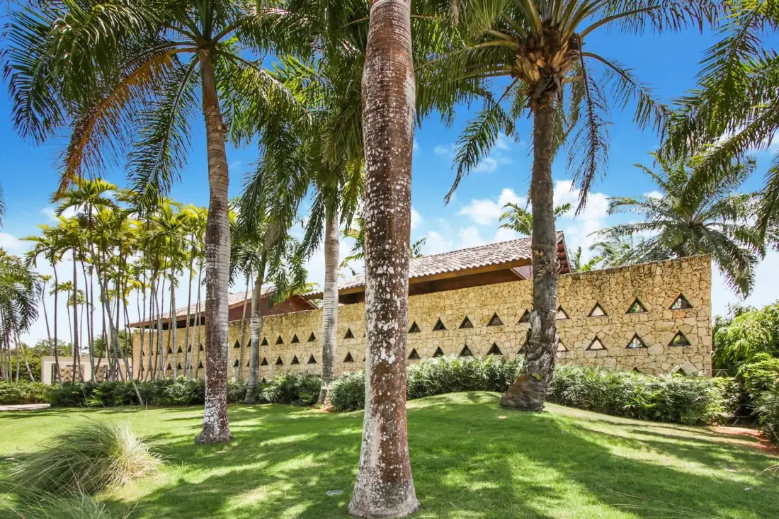 A low stone building stands behind palm trees on a sunny lawn.