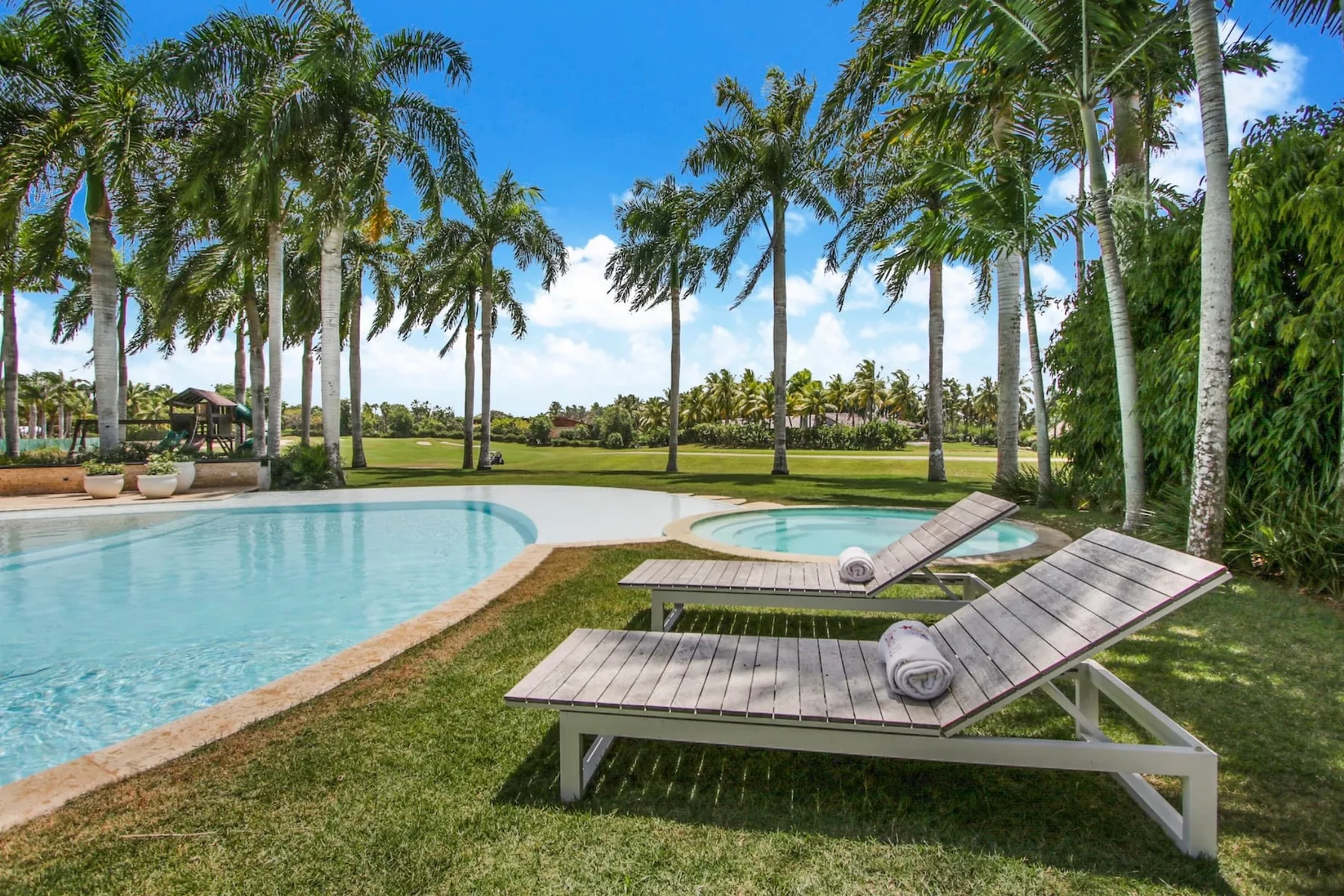 A swimming pool beside two loungers, with palm trees and open lawn in the background.