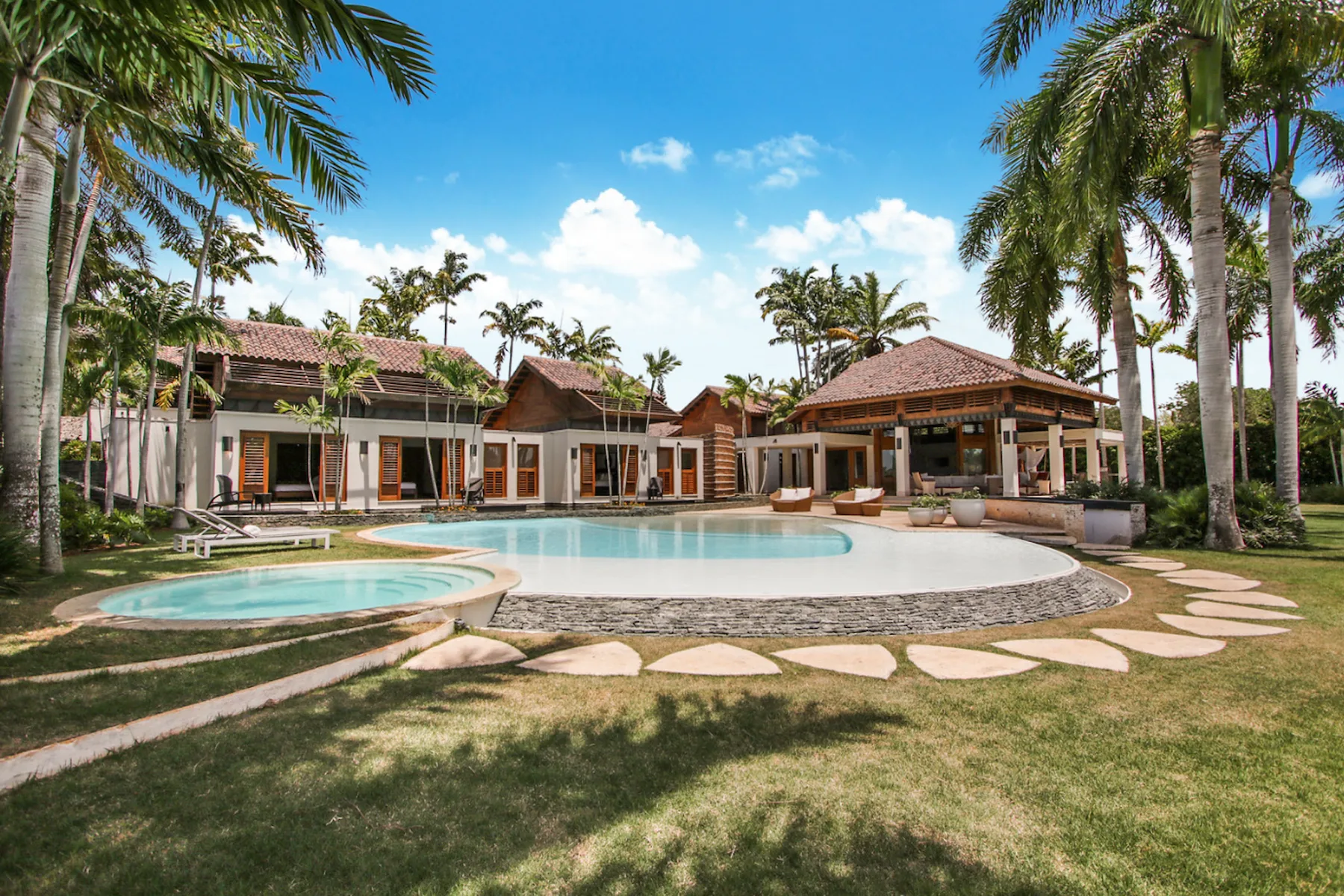 A tropical villa surrounds a swimming pool, with palm trees and a lawn.