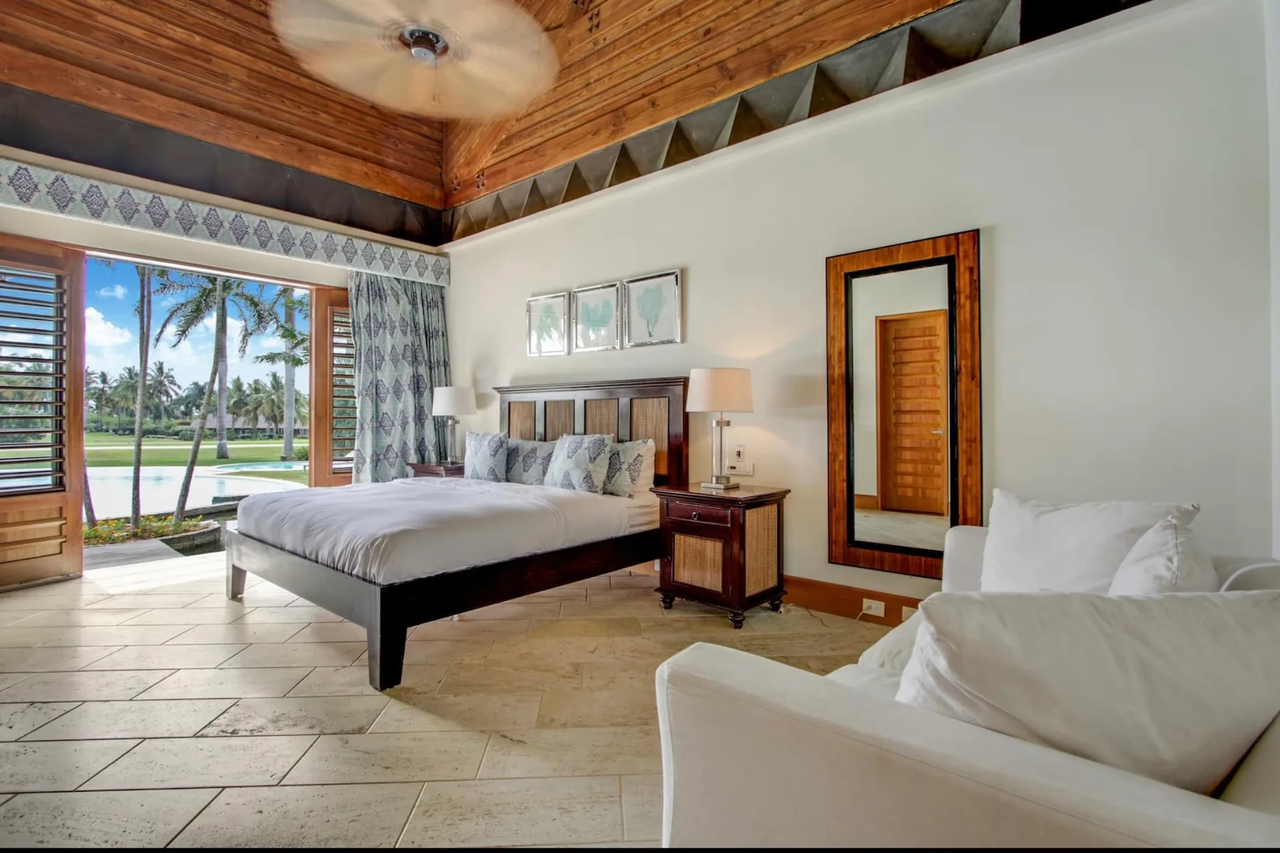 A bedroom with a bed, a seating area, and open doors overlooking palm trees.