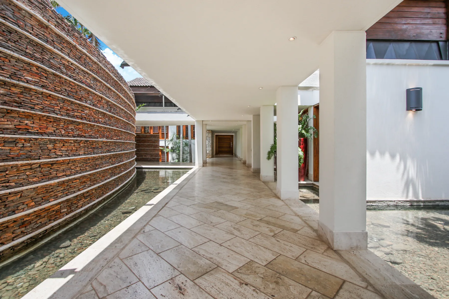 A covered walkway with tiled flooring runs alongside a curved stone wall and water feature.