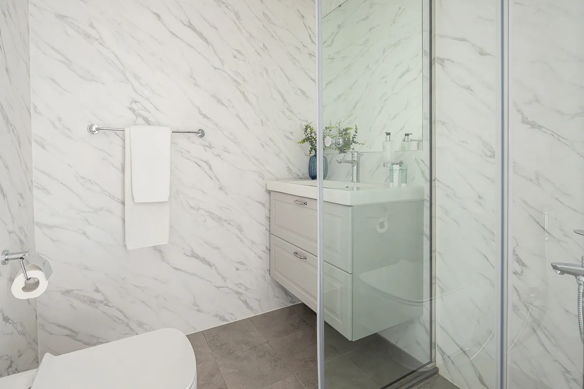 Un baño con ducha de vidrio, tocador y paredes blancas con vetas.