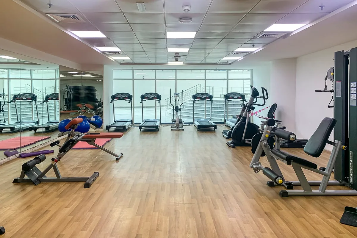 Un gimnasio con máquinas de ejercicio y cintas de correr alrededor de un espacio abierto con suelo de madera.