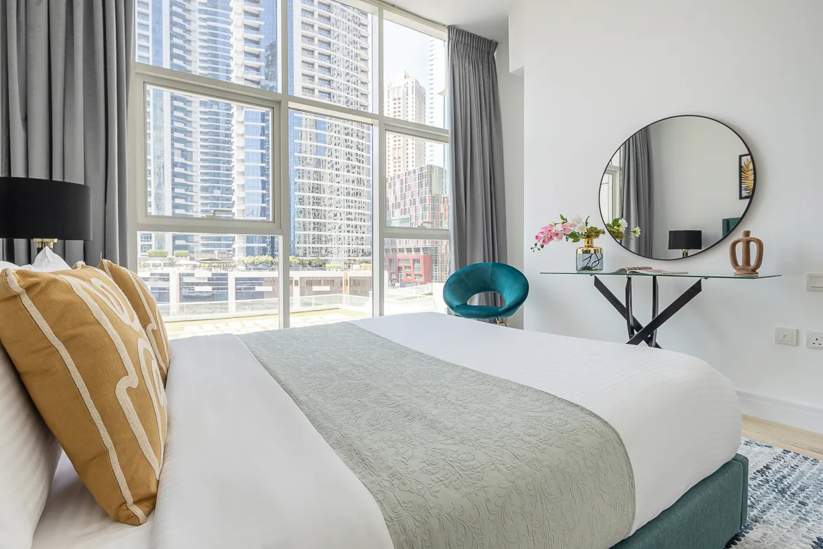 Un dormitorio luminoso con una cama tendida junto a grandes ventanas con vistas a edificios de la ciudad.