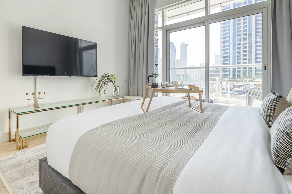 Un dormitorio luminoso con una cama bien arreglada y grandes ventanas con vistas a edificios de la ciudad.