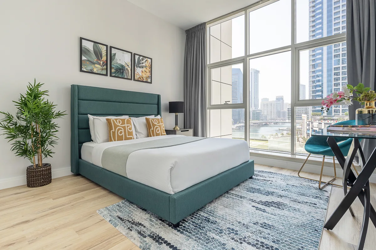 Un dormitorio con una cama color verde azulado junto a grandes ventanas con vistas a la ciudad.