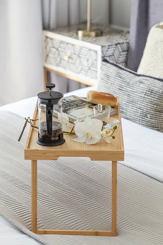 Una pequeña bandeja de desayuno de madera con café y flores está sobre una cama.