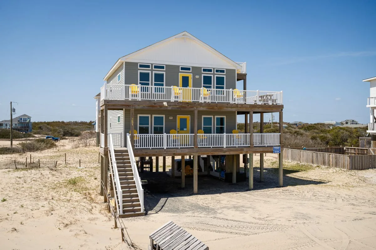 Una casa elevada junto a la costa tiene dos niveles de porches con barandillas blancas y terreno arenoso.