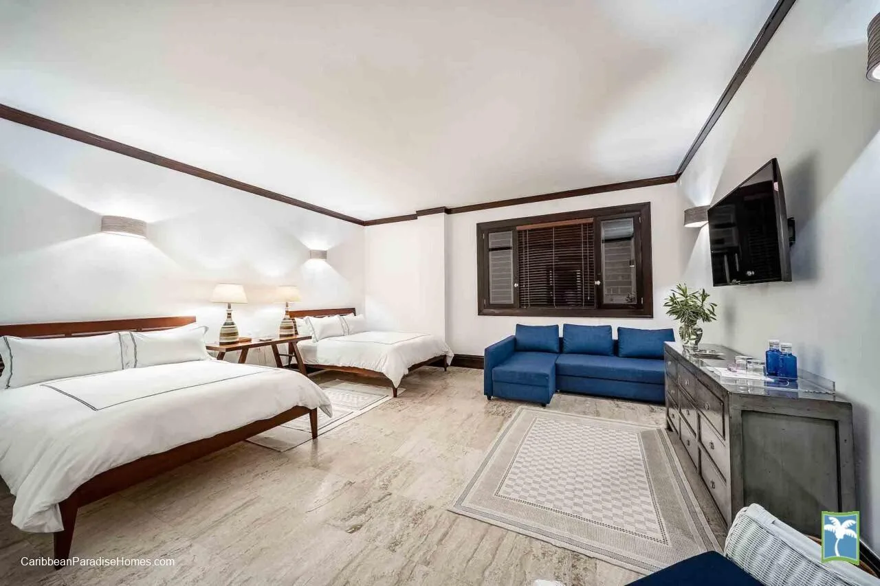 Un dormitorio luminoso con dos camas, una zona de estar azul y un televisor en la pared.
