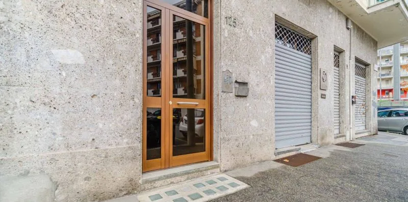 Entrada de un edificio con una puerta de vidrio y marco de madera junto a persianas metálicas cerradas.