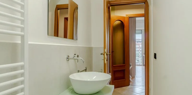 Un baño con un lavabo blanco sobre encimera y una puerta de madera hacia una habitación luminosa.